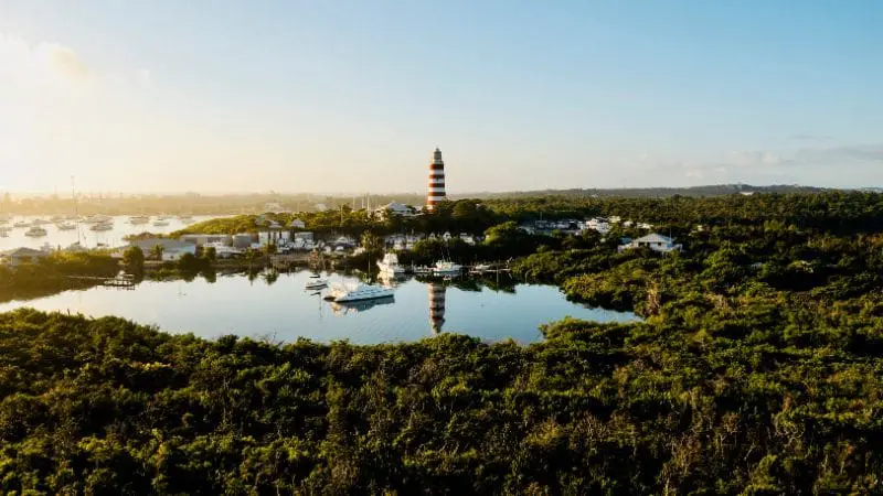 Aerial view of Elbow Cay, Bahamas, featuring a distinctive striped lighthouse amidst lush greenery and marina with boats.