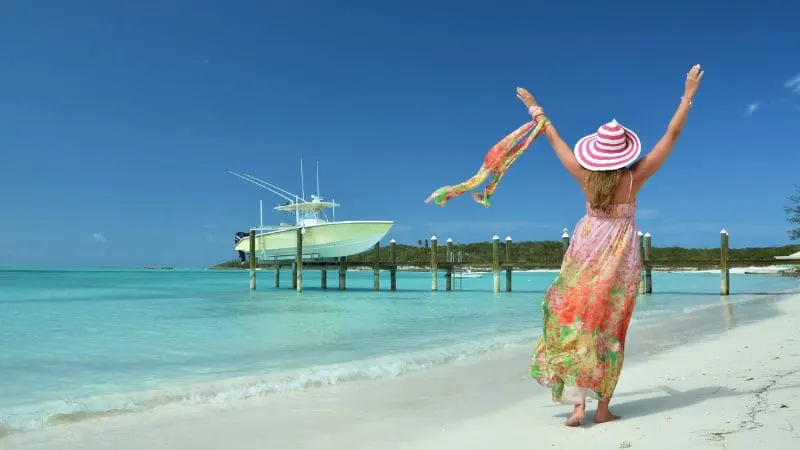 Woman in a floral dress and striped hat enjoying the pristine beach of Exuma, Bahamas, with a docked boat in the background.