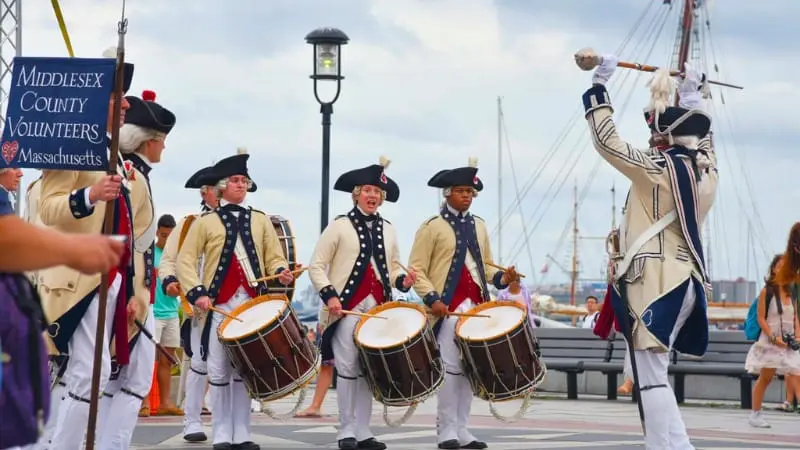 Colonial reenactors march with drums, lively 4th of July parade at a historic destination.