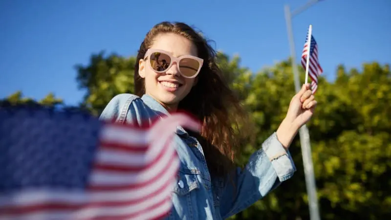 Smiling woman waves flag outdoors, joyful 4th of July scene.