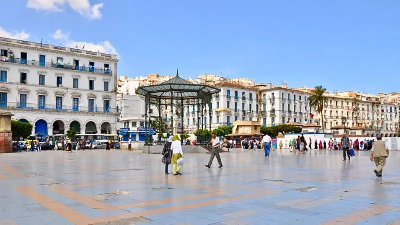 Residents and tourists walk through a square in Casbah Algiers, surrounded by historic buildings.