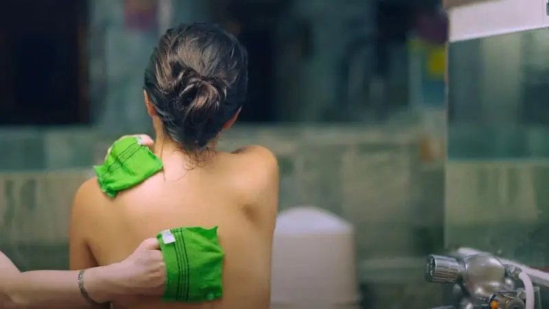 A woman wearing a green towel prepares for a scrub treatment at a Korean spa.