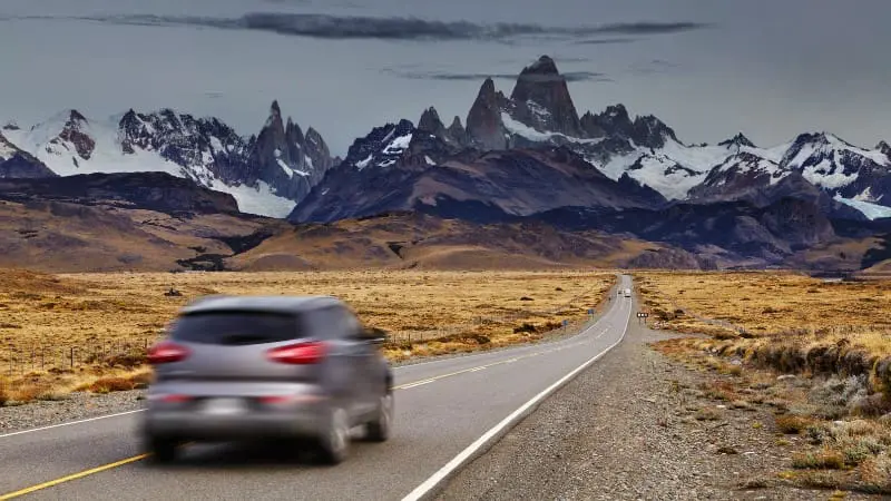 Car drives towards Mount Fitz Roy on a long road through dry Patagonian plains.