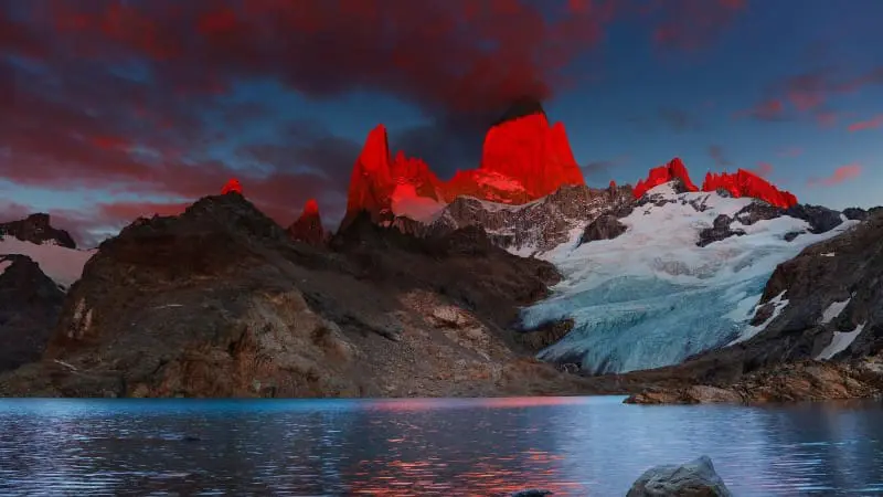 Sunrise turns Mount Fitz Roy peaks red as they reflect in the calm blue lake.