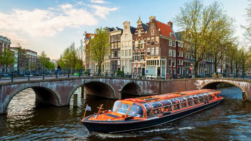 A canal boat cruising under a bridge, surrounded by historic Amsterdam houses.