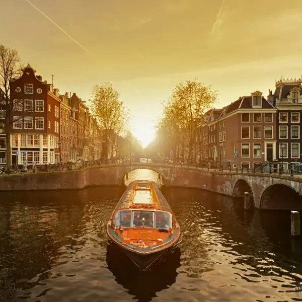 An orange canal boat cruising under a bridge during a golden Amsterdam sunset.
