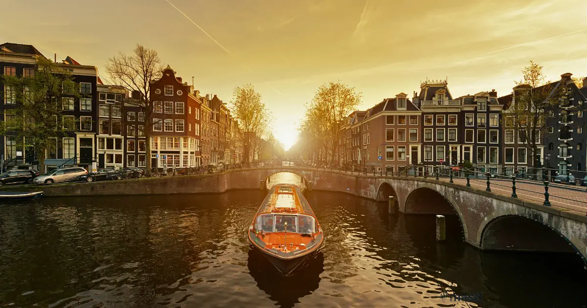 An orange canal boat cruising under a bridge during a golden Amsterdam sunset.