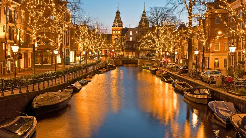Amsterdam canal glowing with festive lights and cozy boats under a stunning evening sky.
