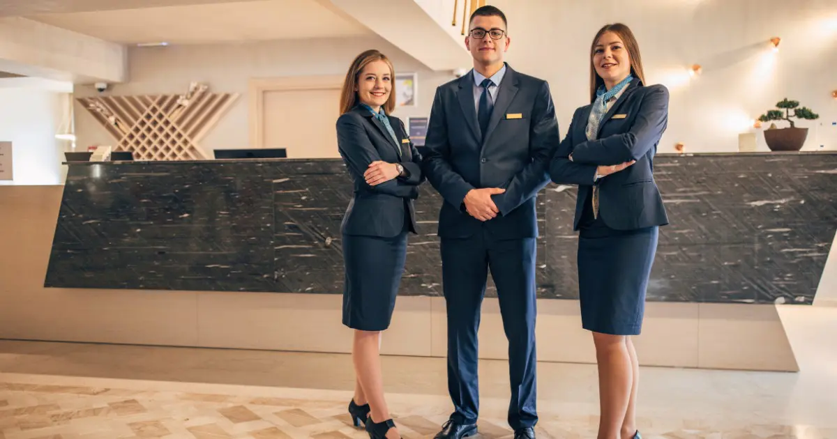 Three hotel staff members in suits welcome guests at marble reception desk.