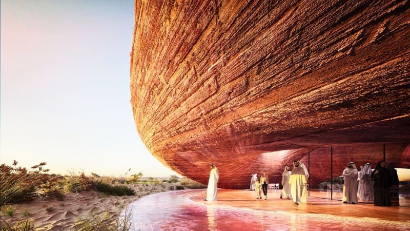 A group of people in traditional clothing stands under a large, modern, rock-like structure at Al Wathba Wetland Reserve, surrounded by a sandy landscape and grass.