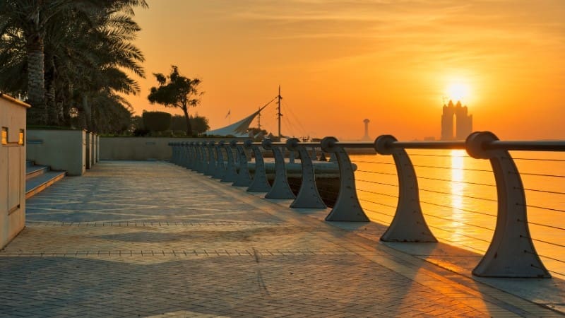 A serene sunset view at Corniche Beach, Abu Dhabi, with a paved walkway, modern railings, palm trees, and calm waters.