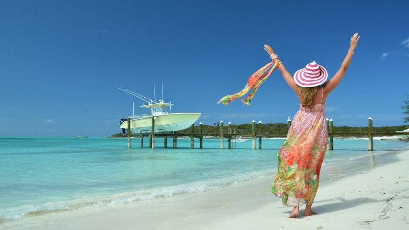 Woman in a floral dress and striped hat enjoying the pristine beach of Exuma, Bahamas, with a docked boat in the background.