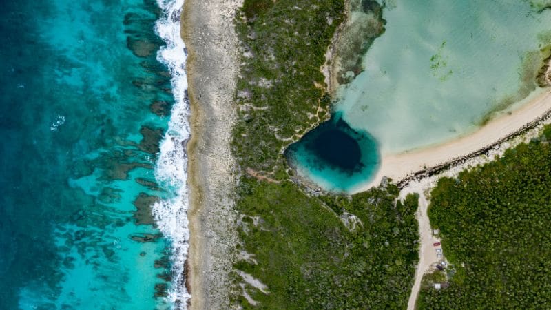 Aerial view of Long Island, Bahamas, with vivid turquoise waters, a sandy shoreline, and a distinctive deep blue hole surrounded by greenery.