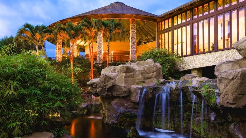 Luxurious Hotel Wailea with tropical garden, waterfall, and lit gazebo at dusk.