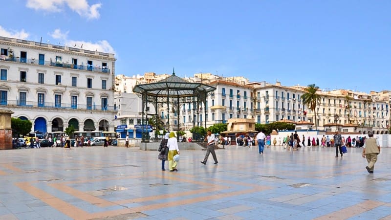 Residents and tourists walk through a square in Casbah Algiers, surrounded by historic buildings.