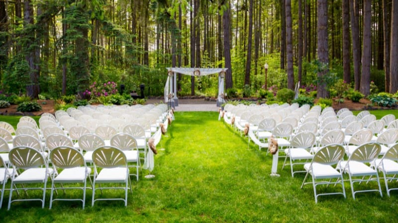 Forest wedding setup in Alabama with white chairs and arch among tall trees.